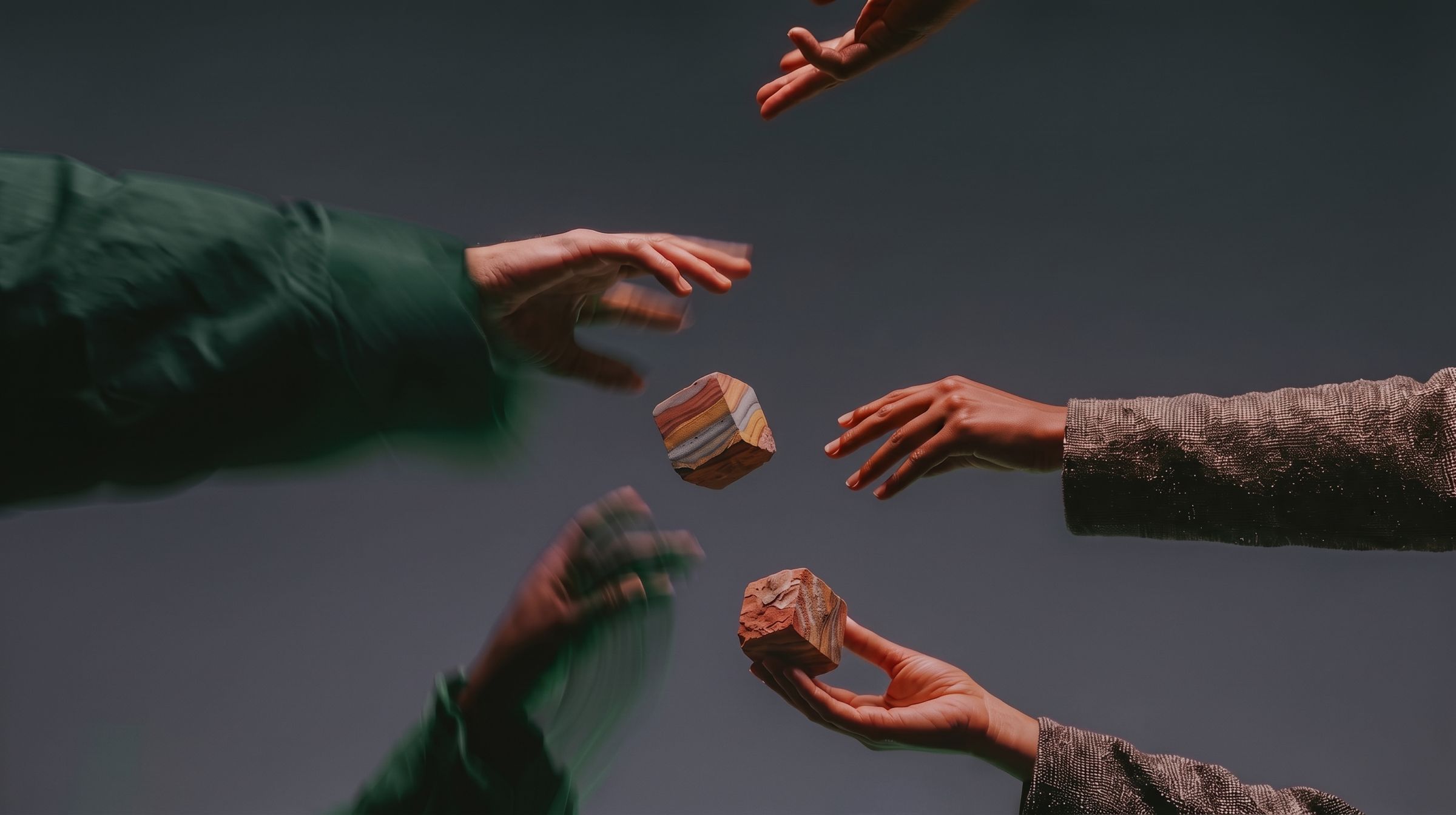 Hands reaching from all sides toward two banded stones in mid-air against a grey backdrop